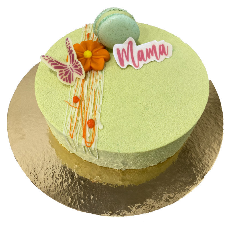 goudkarton met groene taart, met witte en oranje chocoladestrepen, chocoladevlinder met roze vleugels, oranje chocoladebloem, macaron met pistache en chocoladeplak met "mama" erop.